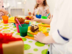 Mesa colorida com bolo, doces e copos, com mãos de crianças alcançando a comida