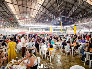 Pessoas comem e bebem em mesas brancas numa feira gastronômica dentro de um mercado coberto.