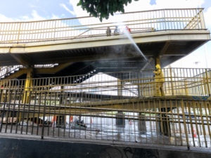 Pessoa em macacão amarelo jateia água sob passarela metálica amarela, com névoa visível.