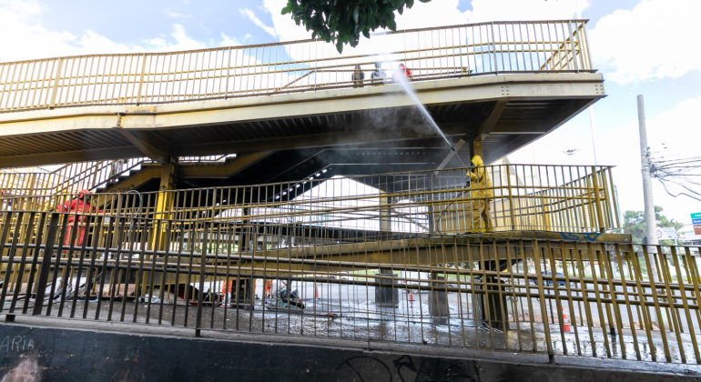 Pessoa em macacão amarelo jateia água sob passarela metálica amarela, com névoa visível.