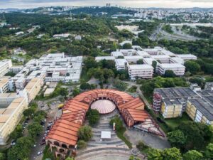 Vista aérea de um campus universitário com edifícios, áreas verdes e um prédio curvo de telha vermelha.
