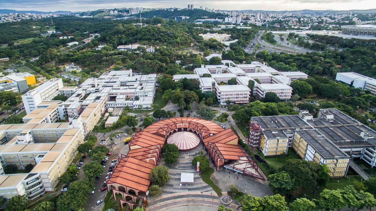 Vista aérea de um campus universitário com edifícios, áreas verdes e um prédio curvo de telha vermelha.