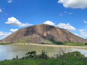 Grande montanha rochosa escura ao lado de um lago calmo sob céu azul com nuvens.