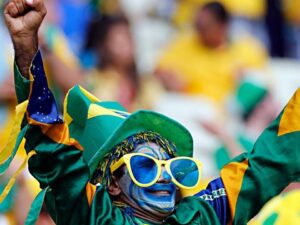 Torcedor com pintura facial, óculos, chapéu e camisa do Brasil, com braços erguidos.