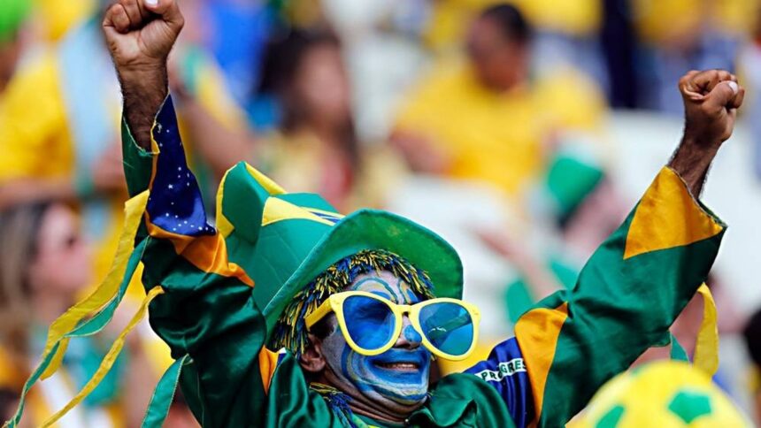 Torcedor com pintura facial, óculos, chapéu e camisa do Brasil, com braços erguidos.