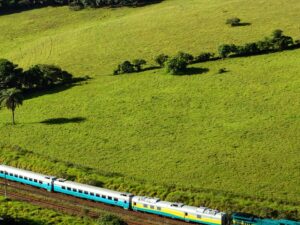 Trem de passageiros multicolorido em trilhos através de um vasto campo verde com árvores e colinas.