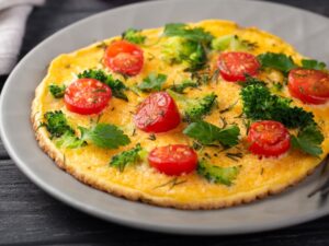Omelete dourado com tomates, brócolis e ervas frescas em prato cinza com talher.