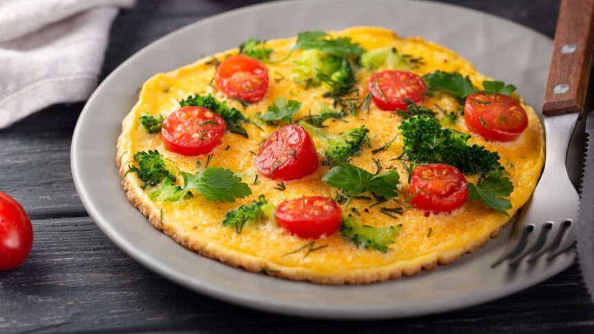 Omelete dourado com tomates, brócolis e ervas frescas em prato cinza com talher.
