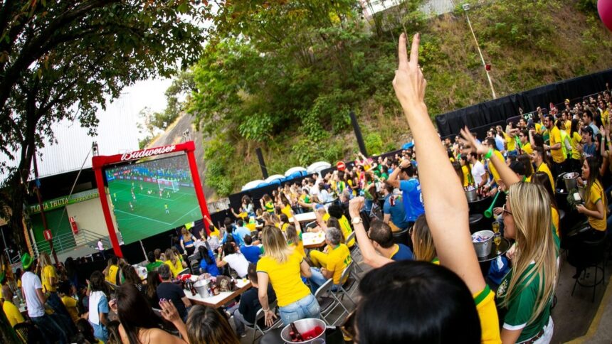 Multidão assiste jogo de futebol em telão Budweiser, pessoas de amarelo/verde torcendo.