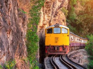 Trem amarelo e vermelho em trilhos curvos próximo a penhascos rochosos e floresta exuberante.