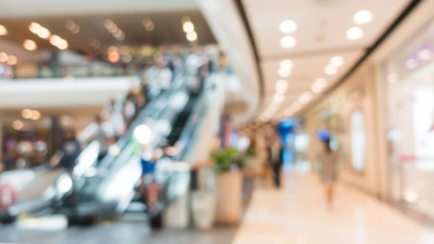 Imagem desfocada de interior de shopping, com escadas rolantes movimentadas e iluminação ambiente.