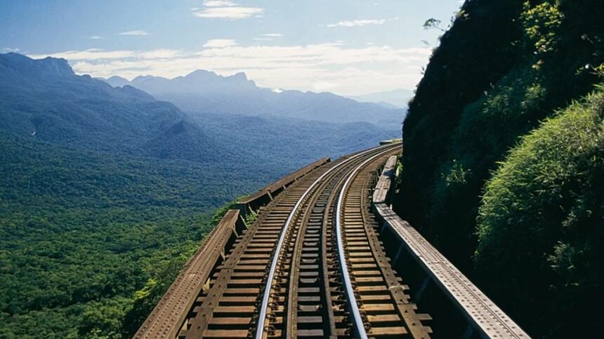 Trilhos de trem em curva, suspensos em encosta montanhosa arborizada, com céu azul e nuvens.