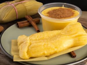 Pamonha, curau com canela, canela em pó e paus em prato verde sobre madeira.