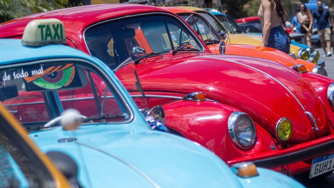 Fila de carros Volkswagen Fusca coloridos, azul-claro com letreiro táxi e vermelho.