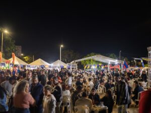 Multidão em evento noturno com tendas iluminadas, pessoas sentadas e em pé, e infláveis coloridos ao fundo.