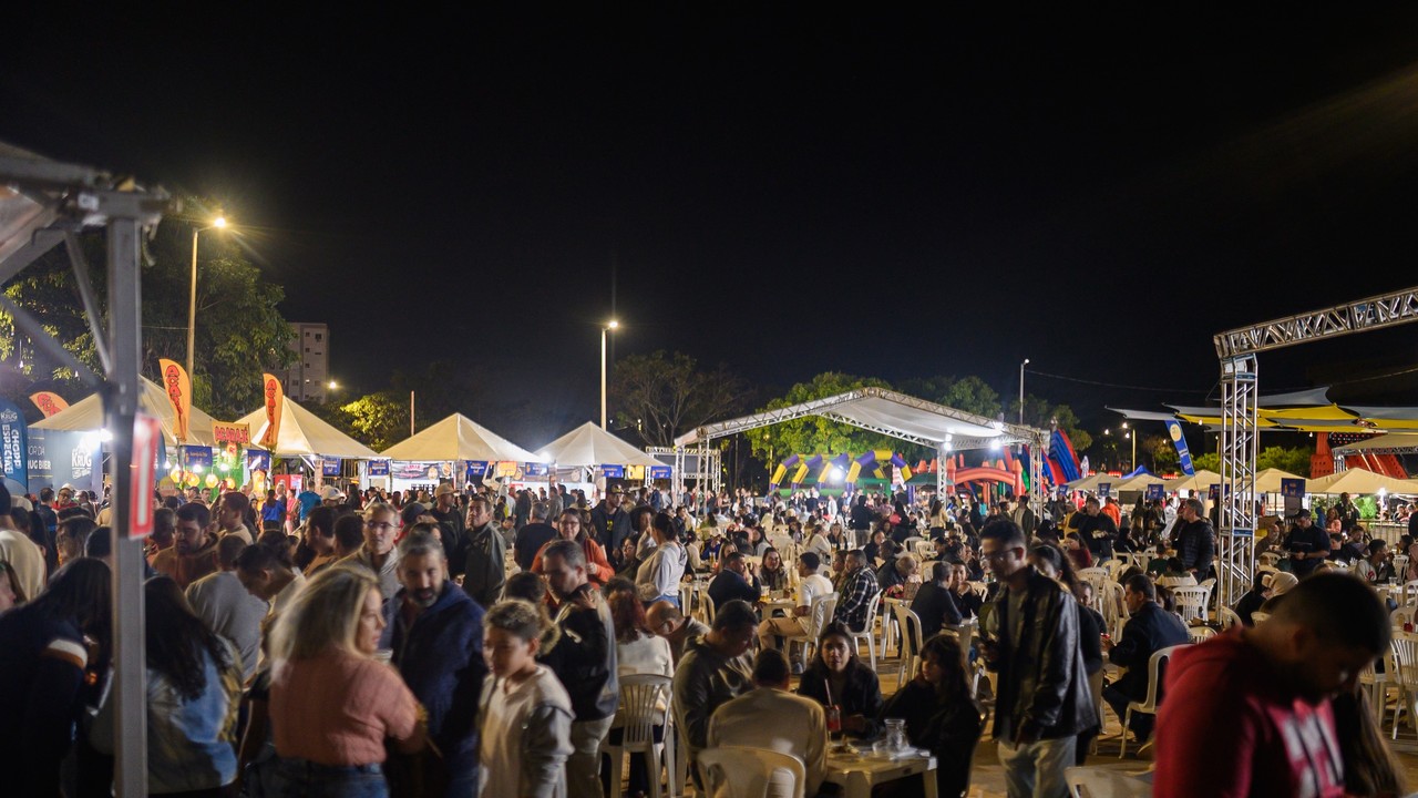 Multidão em evento noturno com tendas iluminadas, pessoas sentadas e em pé, e infláveis coloridos ao fundo.