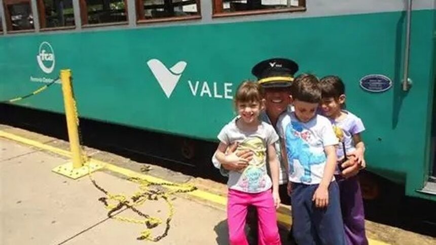 Homem uniformizado sorri abraçando três crianças em frente a um trem verde da Vale.