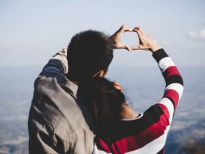 Casal de costas formando coração com as mãos em dia claro, céu azul.