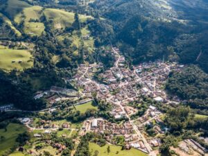 Vista aérea de uma pequena cidade em vale montanhoso, cercada por florestas e campos verdes.