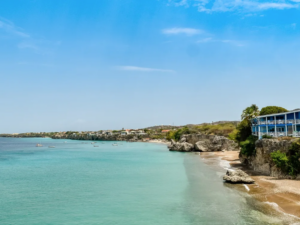 Praia caribenha com água azul-turquesa, falésias rochosas e edifício azul em destaque.