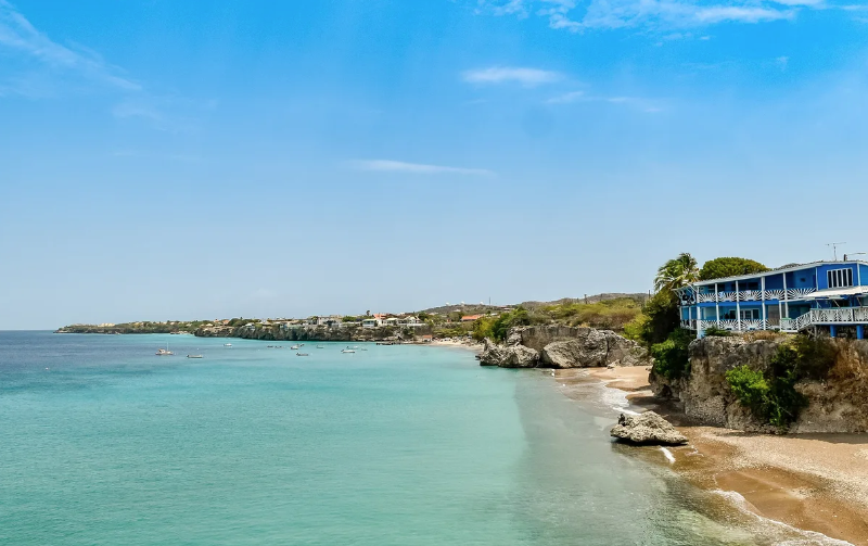 Praia caribenha com água azul-turquesa, falésias rochosas e edifício azul em destaque.