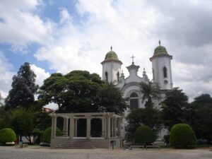 Igreja branca com cúpulas verdes e cruz, junto a um coreto, árvores e céu nublado.