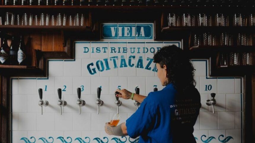 Mulher enche copo de cerveja em bar com torneiras e letreiro "DISTRIBUIDOR GOITACAZI".