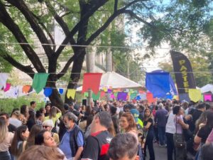 Multidão em praça decorada com bandeirinhas coloridas, com barracas e bandeira 'PASTEL'.