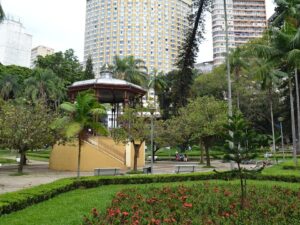 Coreto em parque urbano com árvores, caminhos e prédios altos ao fundo.