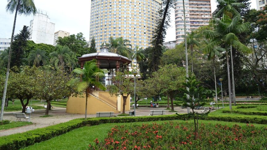 Coreto em parque urbano com árvores, caminhos e prédios altos ao fundo.
