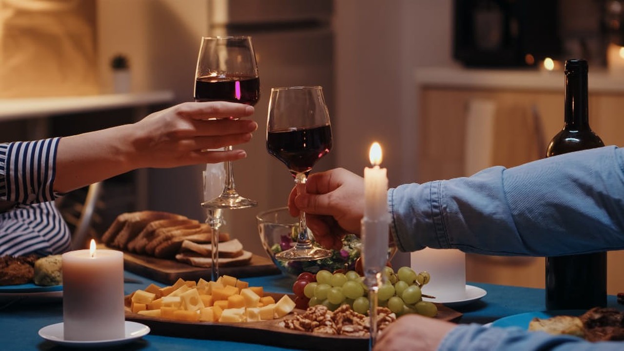 Mãos brindando com taças de vinho tinto, velas acesas e aperitivos em um jantar romântico.