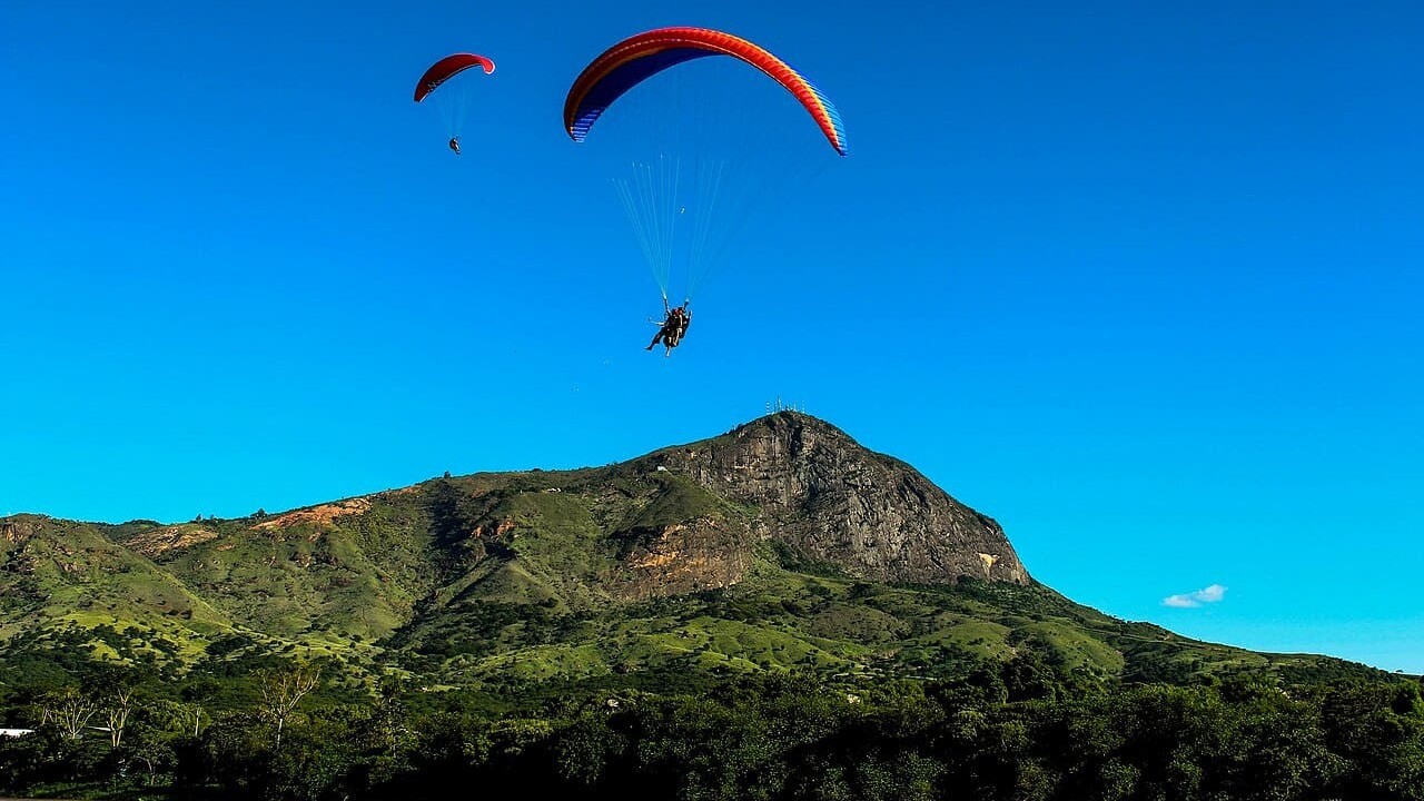 Dois parapentes coloridos voam sobre uma montanha rochosa e verde sob céu azul claro.