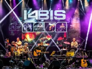 Banda 14 Bis no palco. Músicos com guitarras, teclado, bateria. Telão exibe '14 BIS'.