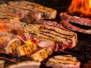 Carne de boi, linguiças e costelas grelhando em uma churrasqueira com chamas fortes.