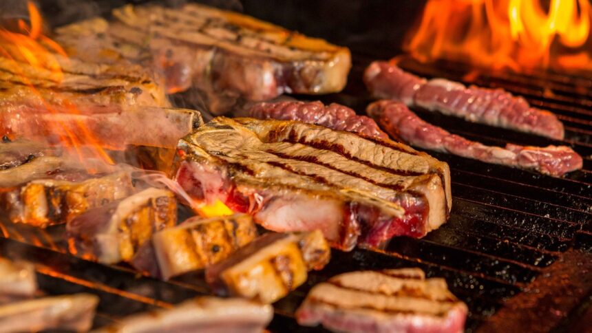 Carne de boi, linguiças e costelas grelhando em uma churrasqueira com chamas fortes.