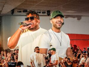 Dois cantores em palco, um com microfone, outro com boné, diante de público animado.