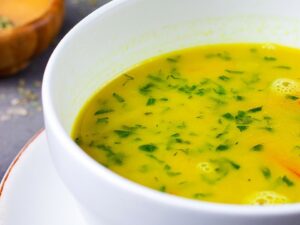 Close-up de tigela de caldo cremoso amarelo com ervas frescas em uma mesa escura.
