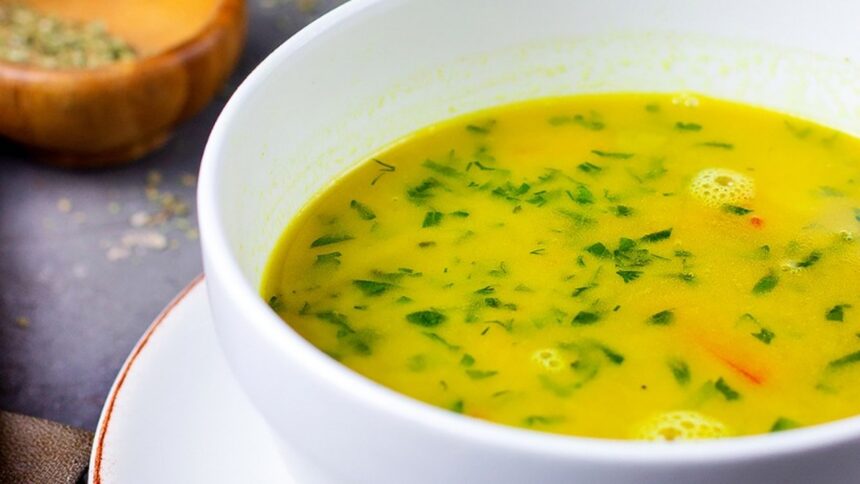 Close-up de tigela de caldo cremoso amarelo com ervas frescas em uma mesa escura.
