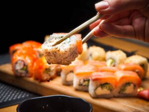 Mão segura sushi com hashi, gota de molho pinga. Bandeja com diversos sushis ao fundo.