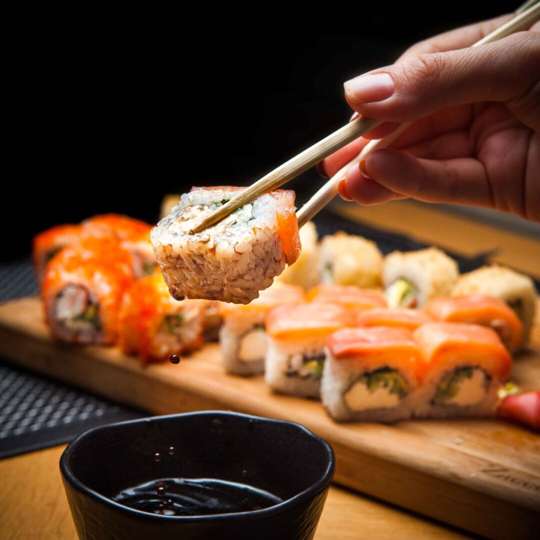 Mão segura sushi com hashi, gota de molho pinga. Bandeja com diversos sushis ao fundo.