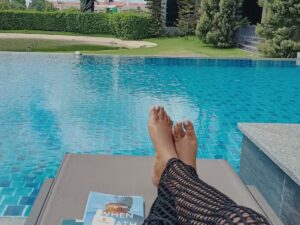 Pés de pessoa relaxando em espreguiçadeira à beira de piscina azul com um livro