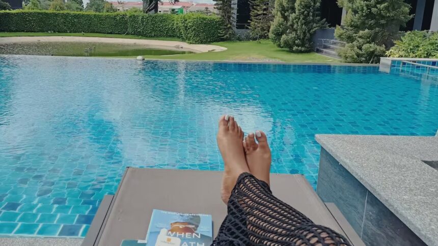 Pés de pessoa relaxando em espreguiçadeira à beira de piscina azul com um livro