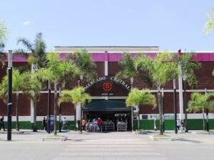 Entrada do Mercado Central de Belo Horizonte com placa "MERCADO CENTRAL DESDE 1929", palmeiras e pessoas.