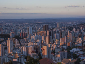 Vista panorâmica da cidade com muitos prédios, alguns iluminados pelo sol poente, e colinas ao fundo.