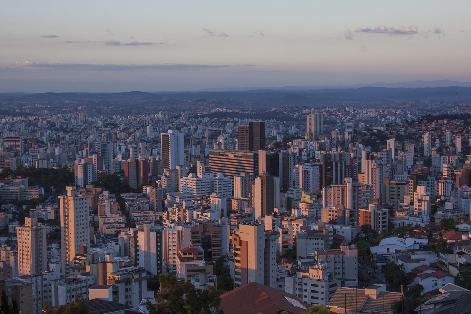 Vista panorâmica da cidade com muitos prédios, alguns iluminados pelo sol poente, e colinas ao fundo.