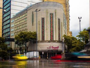 Fachada do Cine Theatro Brasil Vallourec em estilo Art Déco, com rua molhada e veículos em movimento.