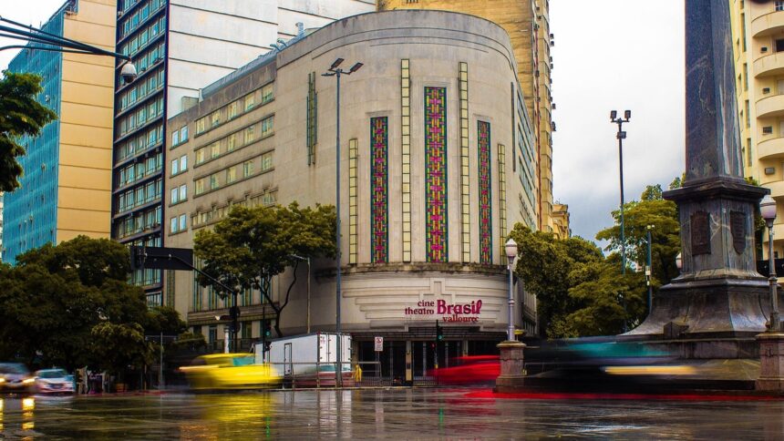 Fachada do Cine Theatro Brasil Vallourec em estilo Art Déco, com rua molhada e veículos em movimento.