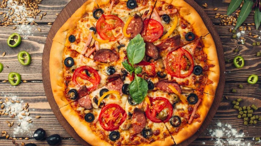 Pizza inteira fatiada com tomate, azeitonas, salame, presunto, pimentões e manjericão na tábua de madeira