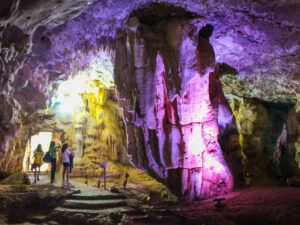 Pessoas exploram interior de gruta com estalactites e estalagmites iluminadas por luzes coloridas