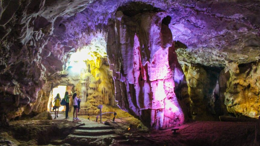 Pessoas exploram interior de gruta com estalactites e estalagmites iluminadas por luzes coloridas